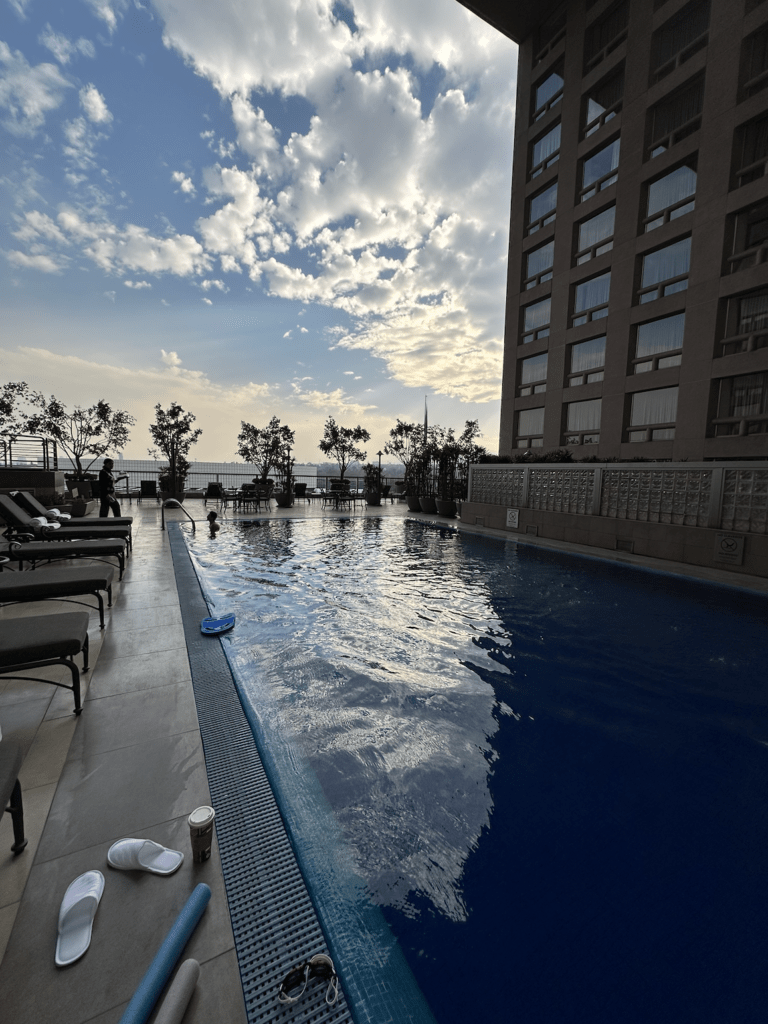 A rooftop swimming pool beside a tall building at sunset, with lounge chairs, slippers, a coffee cup, and pool noodles on the deck—a perfect scene for any travel blog or travel guide featuring unique travel destinations.